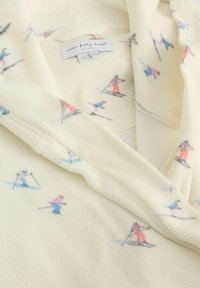 Kremowa tkanina o fakturze z nadrukiem małych kolorowych narciarzy oraz metka odzieżowa z napisem „never fully dressed” wewnątrz kołnierza.