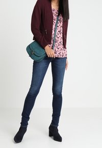 Bordowy kardigan na zamek błyskawiczny na różowej bluzce w kwiaty, do tego ciemnoniebieskie obcisłe jeansy i czarne botki za kostkę. Zielona torebka na ramię.