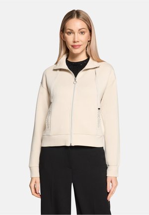 Cream zip-up sweatshirt with a stand collar, front pockets, and decorative accents. Features a fitted design and ribbed cuffs.