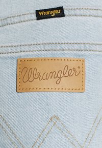 Světlomodré džínové kapsy s hnědou koženou nášivkou a černou látkovou cedulkou, obě označené značkou "Wrangler" vyšitým nápisem.