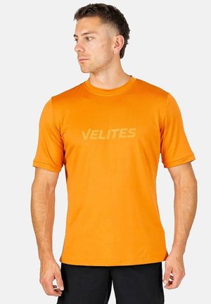 Hombre con camiseta de manga corta de color naranja brillante con el texto "VELITES", mirando hacia la izquierda, de pie contra un fondo liso y claro.