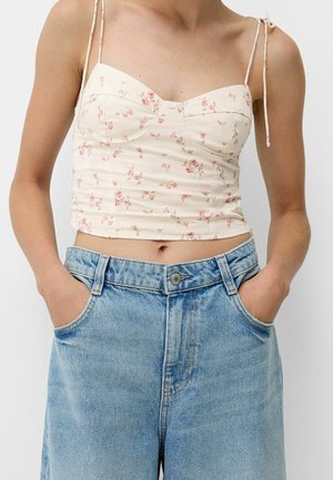 Femme portant un crop top crème à motif floral avec des bretelles spaghetti nouées aux épaules et un jean denim bleu clair taille haute, les mains dans les poches.