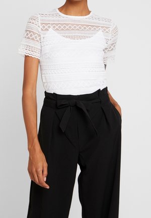 Mujer con una blusa blanca de encaje de manga corta sobre una camiseta blanca, combinada con pantalones negros de talle alto atados con un lazo frontal.