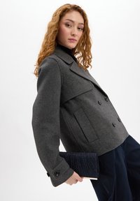 Grijze double-breasted wollen jas met kraag, grote zakken en gebuttonede manchetten, gecombineerd met een marineblauwe geplooide clutch.