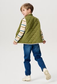 Grüne gesteppte Weste mit hohem Kragen, getragen über einem gestreiften Langarmshirt, kombiniert mit blauen Jeans und hellen Sneakers.
