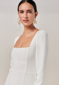 Abito bianco con scollatura quadrata, maniche lunghe a sbuffo e una superficie liscia. Orecchini d'oro a spirale e gioielli a catena fine completano il look.