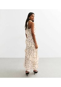 Long, sleeveless dress in a light beige fabric with black polka dots and ruffled layers. Features a halter neck and tied back detail.