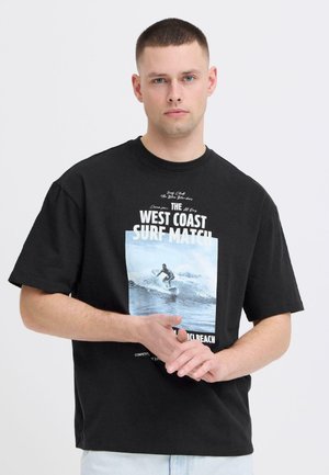 Ung mand med kort hår iført en sort T-shirt med et surfbillede og teksten "West Coast Surf Match."