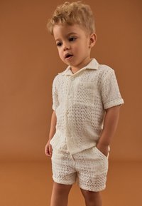 Cămașă albă cu mâneci scurte, texturată, asortată cu pantaloni scurți. Are închidere cu nasturi și detaliu de buzunar. Fotografie realizată pe un fundal maro.