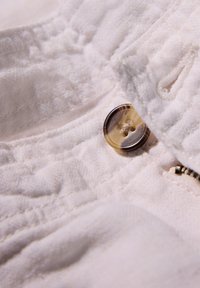 Tessuto bianco con una texture morbida, in primo piano un bottone marrone e un dettaglio della cerniera, che mette in evidenza la cucitura lungo la cucitura.