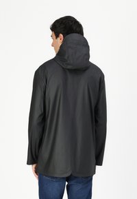 Veste noire imperméable avec capuche, texture lisse et manches longues. Présente une coupe droite et est portée par-dessus un jean bleu.