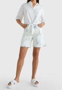 Camicia bianca tagliata con bottoni e nodo frontale, abbinata a pantaloni corti in denim chiaro con motivi blu tie-dye e una cintura bianca.