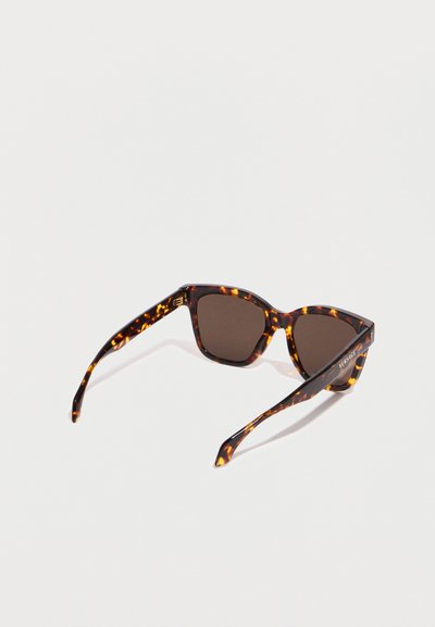 Lunettes de soleil à motif écaille de tortue avec des verres foncés et des montures rectangulaires, inclinées en diagonale sur un fond blanc.