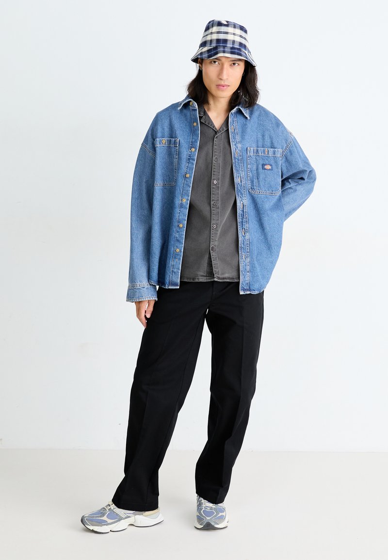 Jeansjacke in blauem Farbton, graues Button-Up-Hemd darunter, schwarze weite Hosen und ein kariertes Bucket Hat. Graue und blaue Muster Sneaker.