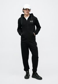 Νεαρός άνδρας φορώντας μαύρη φόρμα EA7 Emporio Armani, άσπρο καπέλο και μαύρα αθλητικά παπούτσια, όρθιος με τα χέρια στις τσέπες της κουκούλας.