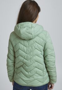 Veste doudoune isolée vert clair avec capuche, présentant des coutures en chevrons, une texture lisse et une silhouette ajustée.