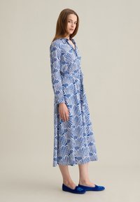 Abito blu e bianco con motivo, maniche lunghe, chiusura con bottoni davanti e cintura in vita. Abbinato a scarpe basse blu con dettagli floreali.