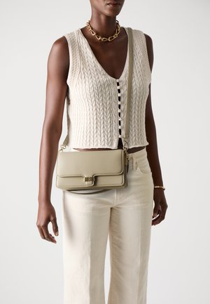 Borsa a tracolla in pelle khaki con finiture dorate, forma rettangolare e chiusura a pattina. Il modello indossa un maglione a trecce colore crema e pantaloni bianchi.