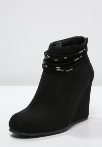 Bottes de cheville noires à talon compensé en daim doux, avec un bout arrondi et plusieurs bandes noires avec des accents métalliques autour de la cheville.