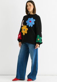 Black knitted sweater with oversized, textured flowers in blue, yellow, red, and green. Paired with wide-leg denim pants and red shoes.