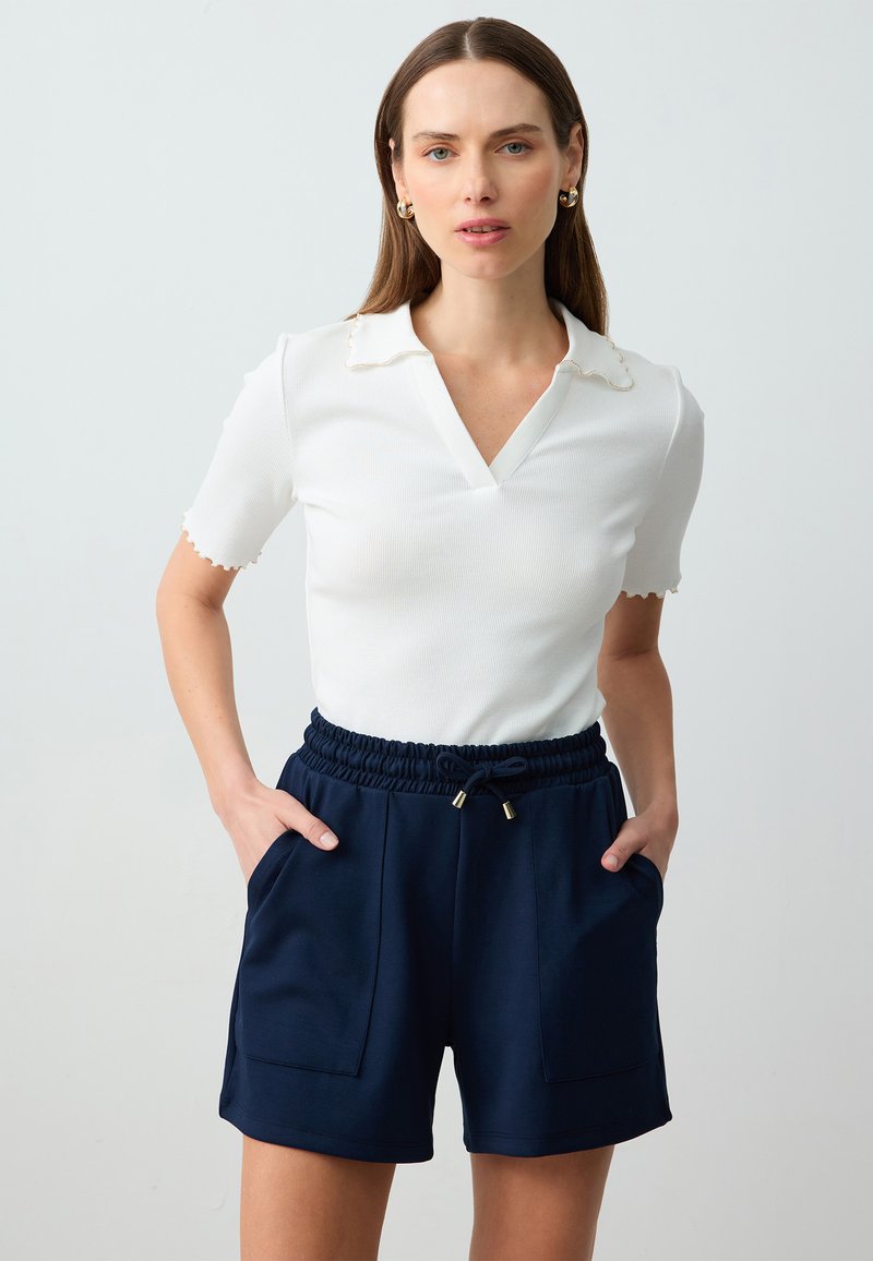 Weißes geripptes Polo-Shirt mit gezacktem Saum, kombiniert mit marineblauen hochgeschnittenen Shorts mit elastischem Bund und zwei Vordertaschen.