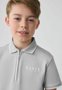 Krátký rukáv, šedá polo košile s límcem na zip, s bílým logem "BAKER BY TED BAKER" na levé části hrudi. Hladká textura.