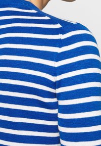 Modrý a bílý pruhovaný pletený svetr s kulatým výstřihem a měkkou texturou, s viditelnými švy a přiléhavým střihem.