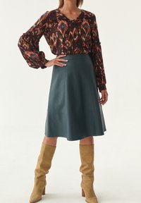 Blusa fluida a fantasia con maniche lunghe, abbinata a una gonna A-line in pelle teal e stivali in suede color sabbia al ginocchio.