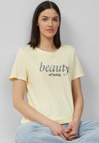 T-shirt jaune clair à manches courtes avec le texte "beauté d'être" en lettres brillantes foncées, fabriqué en tissu doux et texturé.