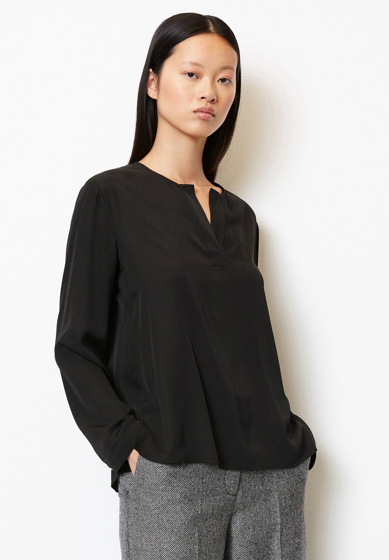 Czarna bluzka o luźnym fasonie, z dekoltem typu popover i długimi rękawami. Miękka tekstura z lekkim drapowaniem. Połączona z wzorzystymi szarymi spodniami.