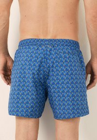 Vista posteriore di un uomo che indossa dei pantaloni da bagno blu con motivo a racchette da tennis e palline, mentre sta in piedi di fronte a un muro bianco.