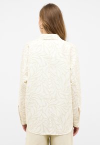 Long-sleeve shirt in light beige fabric with an overlapping leaf pattern, featuring a folded collar and buttoned cuffs. Back view displayed.