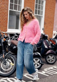Młoda kobieta w luźnych niebieskich dżinsach i różowym, puszystym kardiganie przechodzi obok zaparkowanych motocykli na miejskiej ulicy z budynkami z cegły.