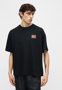 Νέος άνδρας με σγουρά μαλλιά, που φοράει μαύρο T-shirt Nike με κόκκινο λογότυπο, μαύρα σορτς Nike, στέκεται μπροστά σε απλό λευκό φόντο.