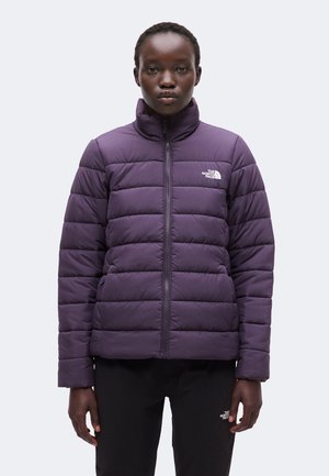 Personne portant une doudoune violette zippée et un pantalon noir, debout face à un fond clair uni.