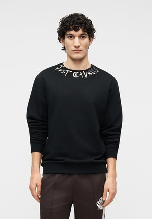 Νεαρός με σγουρά μαλλιά που φοράει μαύρο φούτερ με λευκό κείμενο στο γιακά και σκούρο παντελόνι με λευκό έμβλημα.