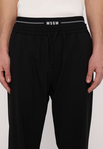 Black jogger pants with an elastic waistband and drawstring. Features white brand logo "MSGM" and striped detailing on the waistband.