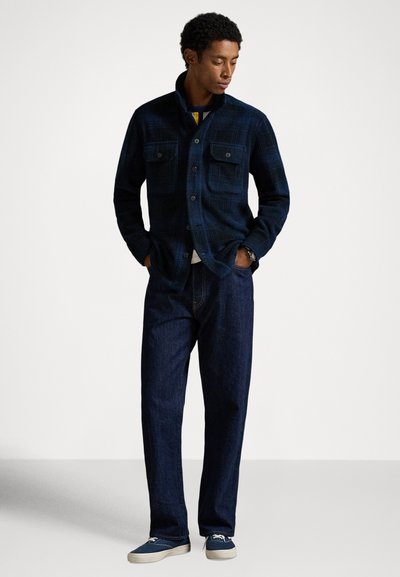 Čovjek stoji, nosi tamnoplavu košulju na kopčanje s kariranim uzorkom, tamne traperice, plave tenisice, ruke u džepovima, gleda dolje u stranu na jednoličnoj pozadini.