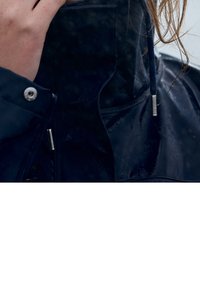 Veste imperméable bleu marine avec col montant, éléments argentés et cordons de serrage réglables ; le matériau semble brillant avec des gouttes d'eau.