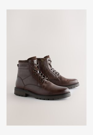 Ruskeat nahkasaappaat, joissa on sileä pinta, nauhallinen edestä ja sivuvetoketju. Vankka kumipohja, jossa uritettu pito. Korkeus keskivartaloon.