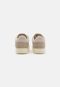 Beige suede sneakers med en glatt tekstur, med avrundet tå og kontrasterende kremfargede gummisåler. Bakre pull-tab for enkel påkledning.