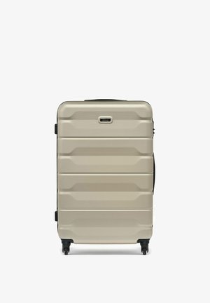 Twarda walizka w kolorze beżowym z poziomymi żłobieniami, czterema czarnymi kółkami obrotowymi, bocznymi i górnymi uchwytami oraz małą tabliczką z logo marki.