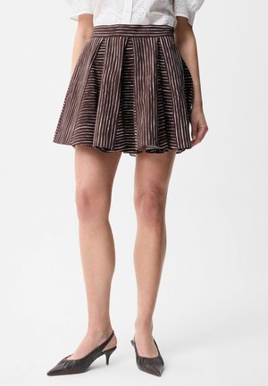 Femme portant une mini-jupe plissée marron avec des rayures verticales blanches, un chemisier blanc à œillets et des chaussures slingback noires à petits talons.