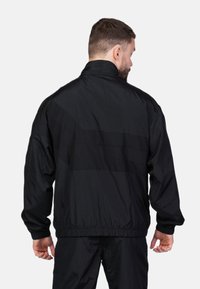 Schwarze Windjacke mit hohem Kragen, elastischen Ärmelbündchen und Bund. Sie verfügt über einen strukturierten Stoff und horizontale Nähte über den Rücken.
