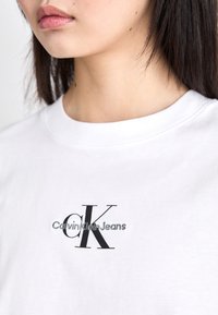 Detailní záběr na osobu v bílém tričku s černým logem "cK Calvin Klein Jeans" vytištěným na prsou.