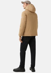 Mann trägt eine beigefarbene Kapuzenjacke, schwarze Jeans, schwarze Schuhe und eine beigefarbene Mütze, steht seitlich vor einem weißen Hintergrund.