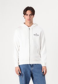 Valge lukuga kapuutsiga sweatshirt, millel on reguleeritav kapuuts ja eesmine taskud. Esiküljel on tagasihoidlik logo, valmistatud pehmest kangast. Kombineeritud siniste teksadega.