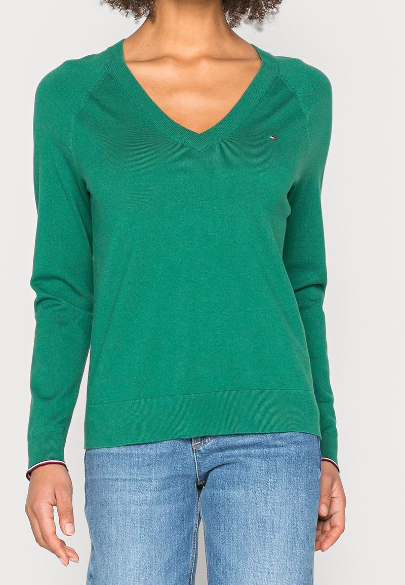 Femme portant un pull vert à col en V avec des manches longues et un jean bleu clair, debout devant un fond uni.