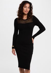 Robe noire côtelée avec des manches longues, présentant un panneau en maille rayée transparente au niveau du col. Design ajusté, longueur genoux.