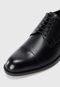Scarpe di pelle nera con punta a cappello e chiusura con lacci, caratterizzate da una punta liscia e una tomaia in pelle texture.